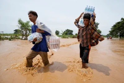 People wading through flood water in Naypyidaw. They are carrying supplies, One man is balancing a pack of water bottles on his head. The flood water is orangey-brown