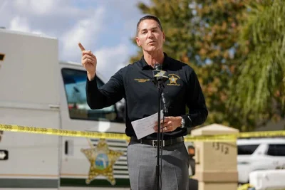 Sheriff Chad Chronister, wearing a black shirt and gray pants, raises his right hand while standing in front of a microphone.