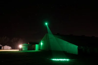 A green laser beam on top of a barn projects a green circle onto the ground at night.