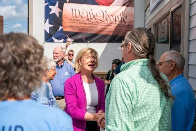 Senator Tammy Baldwin, wearing a magenta sweater over a white T-shirt, shaking hands with a person in a white and green checked shirt. 