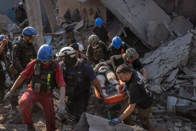 A group of men, most wearing helmets and some wearing camouflage, lifting a person from a pile of rubble.