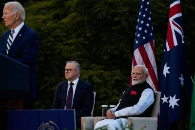 President Biden speaks while two men, one bearded, sit behind him. The Australian and American flags are visible behind the men.