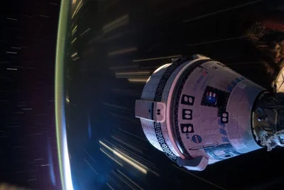 A long-exposure photograph shows the Boeing Starliner spacecraft docked with the International Space Station. It's nighttime on Earth and it shows bright streaks from cities zooming by.