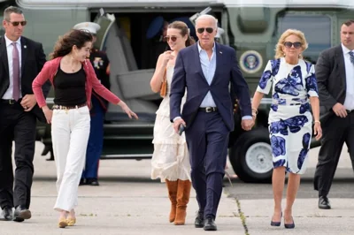 Joe Biden with his wife and other family members. They are walking away from a helicopter. He is wearing a blue suit and aviator sunglasses. Jill biden is dressed in a blue and white dress.