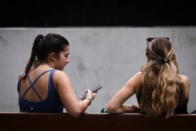 Two adolescent girls sitting on a bench, with one holding a cellphone, seen from behind.