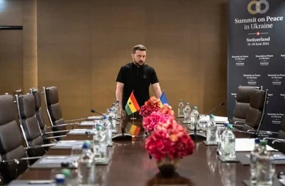 President Volodymyr Zelensky of Ukraine standing at the head of a long table lined with empty chairs. To the right is a sign reading “Summit on Peace in Ukraine.”