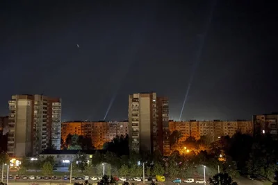 Beams of light rise into the night sky, with buildings and parked cars in the foreground.