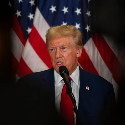 Former President Donald J. Trump speaks into a microphone in front of an American flag. 