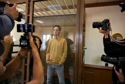 a man in a sweatshirt and jeans stands behind glass as photographers take pictures