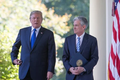 President Donald J. Trump in 2017 walking outdoors with Jerome Powell, who is holding a blue folder, with an American flag next to them and trees in the background.