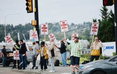 A group of people with signs saying "On strike against Boeing" wave at a car on the side of a road.