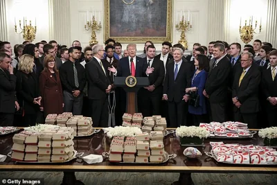 Then-President Donald Trump speaks behind a table full of McDonald's hamburgers, Chick fil-a sandwiches and other fast food as he welcomes the 2018 Football Division I FCS champs North Dakota State Bison in the Diplomatic Room of the White House on March 4, 2019 in Washington, DC COVID protocols impacted kitchens at the time