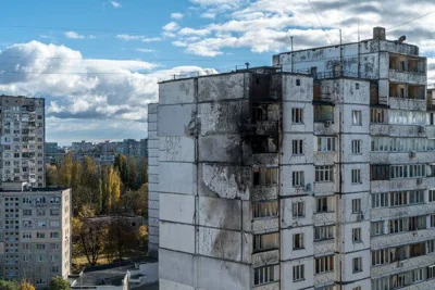 A damaged apartment building with a blackened facade is shown against a clear sky and urban skyline.