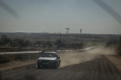 A car drives along a road, sending dust into the air. On the horizon, plumes of smoke can be seen.