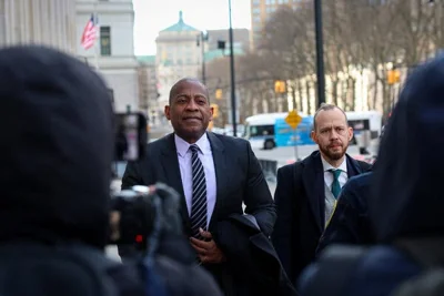 Carlos Watson, wearing a dark suit, light shirt and striped tie, walks on the street next to another man in a suit and tie.