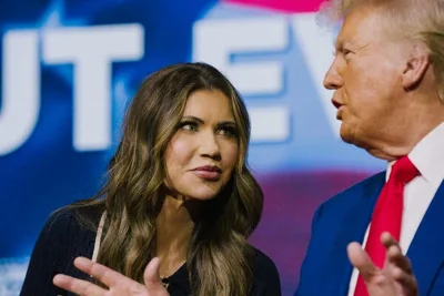 Kristi Noem listens as Donald J. Trump gestures and speaks, with large lettering visible behind them. 