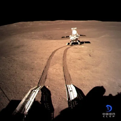 A view of a lunar rover on the surface of the moon from the lander, with two tracks in the lunar dirt made by the rover, a short distance from the lander.