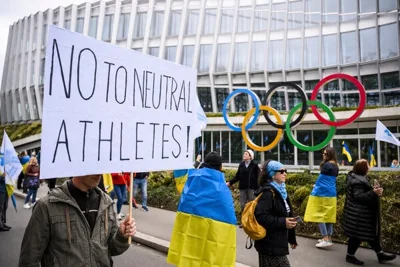 The Olympic logo stands outside of a building as people wearing Ukrainian flags protest. One person holds a sign that reads “No to Neutral Athletes.”