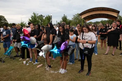 A group of young people stand on a grass lawn with their heads bowed. Two people are holding star shaped balloons.