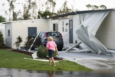 A woman walks through a flooded lawn past severe exterior damage to the roof and siding of an Osprey home