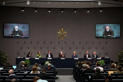 Seven people sitting behind a long table, with two flat-screen televisions on the wall behind them.