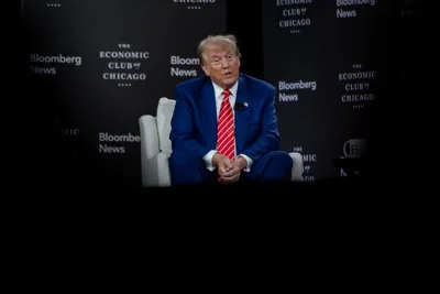 Donald Trump, wearing a blue suit and a red-and-white striped tie, sits in a white armchair onstage. The wall behind him is emblazoned with the logos of the Economic Club of Chicago and Bloomberg News.