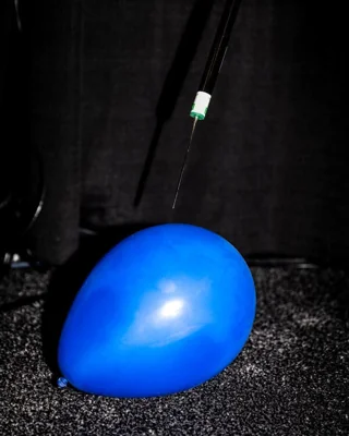 A blue balloon being pierced by a long hypodermic needle.