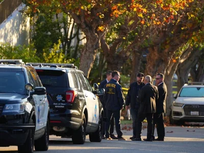 Five law enforcement officers gather in a circle, with two police vehicles visible in the foreground on a fall day, as indicated by trees with their leaves changing color.