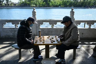 Two men sit at an outdoor table next to a lake playing a game with a grid and large wooden pieces that look like checkers.