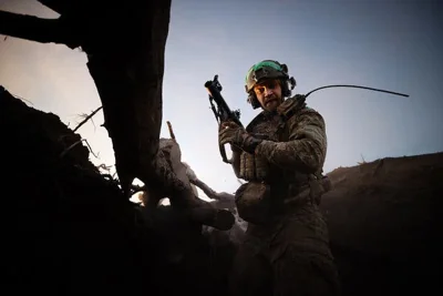 A soldier in a helmet holds a grenade launcher while standing in a trench.