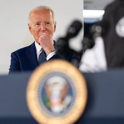 President Biden stands behind a podium with the presidential seal out of focus, with his hand to his mouth.