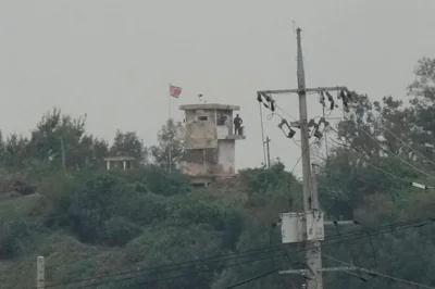 A North Korean soldier standing guard in a watch tower at the border with South Korea. A North Korea flag is flying from the building. There are lots of trees around.