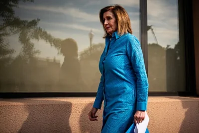 Nancy Pelosi in a blue shirtdress, walking outside near a windowed building.