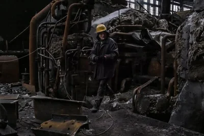 A person in a yellow hard hat walks amid the twisted ruins of a building.