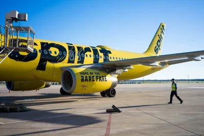 A person in a yellow vest walks behind a yellow jetliner with the words "spirit" and "home of the bare fare” written in black on it.