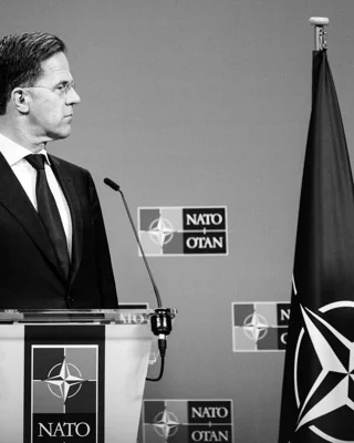 A man in a suit stands at a NATO-branded lectern. 