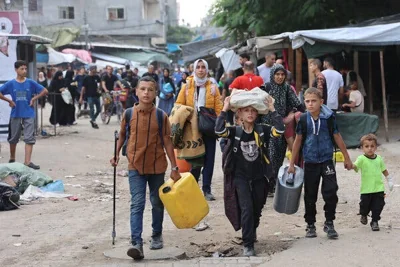 Adults and children carry plastic jugs and other belongings while walking along a road.