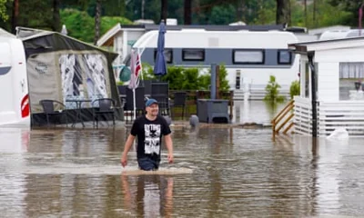 A man walks past camper vans on a flooded campsite: he is smiling despite being thigh-deep in brown, muddy water. The vans and static caravans are swamped with outdoor chairs seen underwater. 