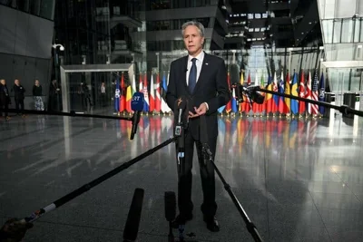 A man in a suit stands in front of a gray and glass office building entrance. Microphones are pointed at him and flags are seen in the background.