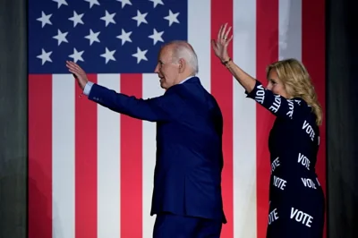 Joe and Jill Biden waving to the crowd as they walk along a stage. There is a large US flag behind them.