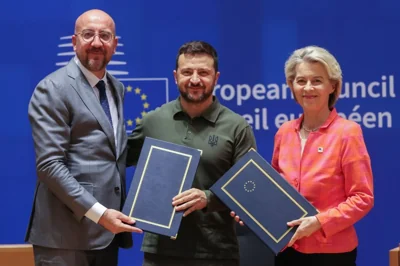 European Council President Charles Michel, European Commission President Ursula von der Leyen and Ukraine's President Volodymyr Zelenskyy. Zelenskyy is in the middle. Michel and von der Leyen are holding blue folders embossed with the EU circle of stars and gold trim. They are all smiling.