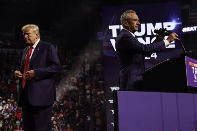 Robert F. Kennedy Jr. stands at a lectern holding the microphone as Donald J. Trump walks away in the background at a campaign rally.