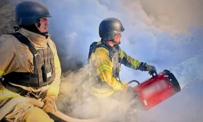 Two men kneeling surrounded by smoke, one throwing a bucket of water on to flames