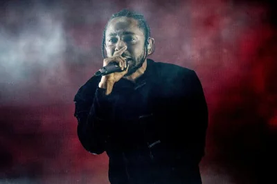 Kendrick Lamar performs on a stage while wearing a black jacket and holding a microphone to his mouth.