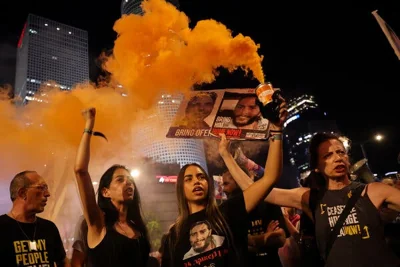 Protesters under a night sky, with skyscrapers in the background, holding signs with photos of hostages that read, “Bring Him Home Now.”