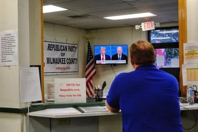 Former President Donald J. Trump and President Biden are displayed on a television as a man in a blue shirt watches.