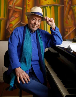 Sergio Mendes, dressed all in blue with a green scarf and a fedora, sits at a piano with his left elbow leaning on the top and smiles.