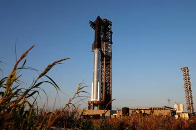 A view from the ground looking up at the extremely tall launch stand with the black Starship spacecraft sitting atop a giant silver cylindrical booster rocket.