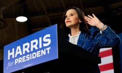 Whitmer gestures behind a lectern on which is written ‘Harris for president’