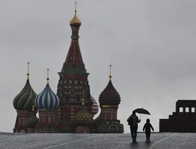 An adult with an umbrella walking with a child is silhouetted against a gray sky on Red Square, with the domes of St. Basil’s Cathedral on the left. 